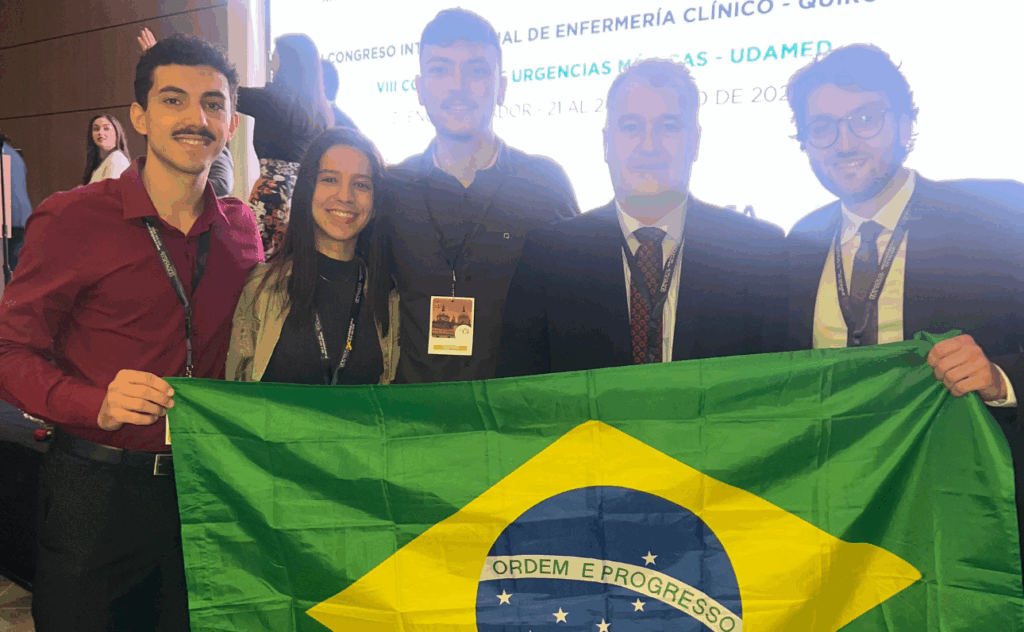 Cinco homens estão em uma sala com janelas amplas e vista para uma cidade ao fundo. Estão sorrindo e posando para a foto, próximos a uma mesa com objetos como celular, óculos, bloco de anotações e um notebook. Os dois homens à direita seguram camisetas brancas com detalhes em azul e amarelo, que exibem bandeiras do Brasil e do Equador, além do texto "Yellow Wave 2025". Os dois homens à esquerda usam roupas casuais (camisetas claras e calças escuras), enquanto os três à direita estão com camisas sociais. Uma cadeira ao lado exibe um jaleco branco com o nome “Dr. […] Cirugía”. O ambiente sugere uma reunião profissional ou preparativo para um evento futuro.