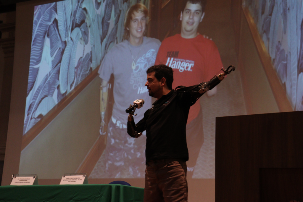 Convidado de honra do WCTEL, o economista e empresário Pedro Pimenta compartilhou sua jornada de superação em palestra emocionante. Aos 19 anos, contraiu uma forma grave de bacteremia que lhe deixou com menos de 1% de chance de sobrevivência e resultou na amputação dos quatro membros. Na imagem, Pedro segura o microfone com um dos braços protéticos enquanto aponta, com o outro braço protético, a imagem no telão, em que aparece uma foto sua ao lado do amigo Cameron Blake.