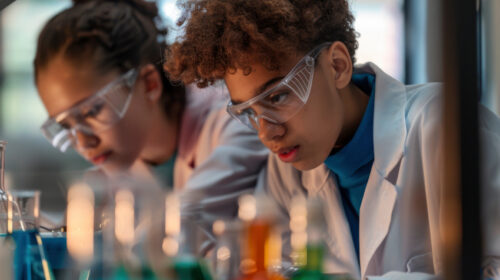 Dois jovens estudantes, usando jalecos brancos e óculos de proteção, estão concentrados em um experimento de laboratório. Eles observam atentamente frascos com líquidos coloridos dispostos à frente, em tons de azul, verde, laranja e amarelo. O ambiente é bem iluminado e transmite uma atmosfera de curiosidade científica e aprendizado prático.
