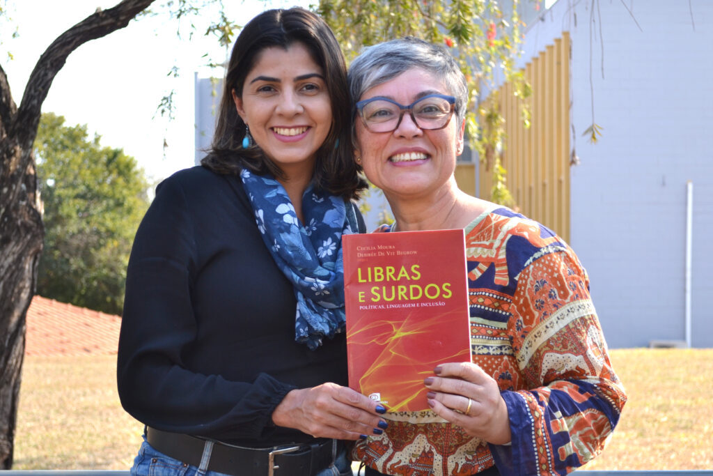 Duas mulheres sorridentes posam juntas ao ar livre segurando o livro "Libras e Surdos: Políticas, Linguagem e Inclusão". A mulher à esquerda veste blusa preta e cachecol azul com estampa floral; a da direita, de cabelos grisalhos curtos, usa óculos e uma blusa estampada colorida. Ambas olham para a câmera.