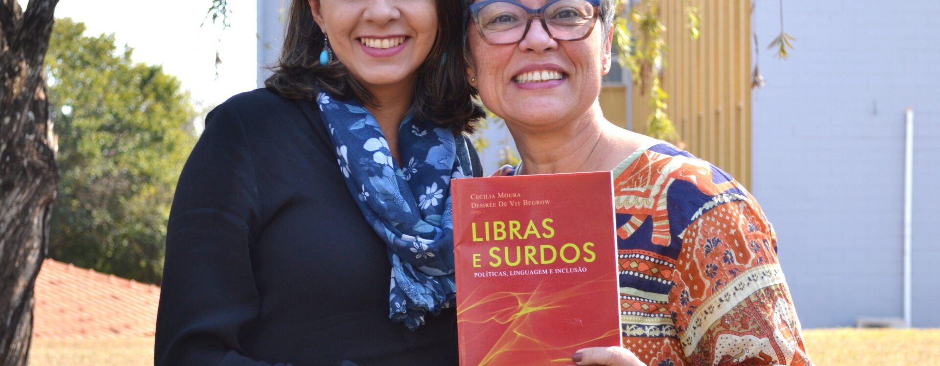 Duas mulheres sorridentes posam juntas ao ar livre segurando o livro "Libras e Surdos: Políticas, Linguagem e Inclusão". A mulher à esquerda veste blusa preta e cachecol azul com estampa floral; a da direita, de cabelos grisalhos curtos, usa óculos e uma blusa estampada colorida. Ambas olham para a câmera.