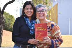 Duas mulheres sorridentes posam juntas ao ar livre segurando o livro "Libras e Surdos: Políticas, Linguagem e Inclusão". A mulher à esquerda veste blusa preta e cachecol azul com estampa floral; a da direita, de cabelos grisalhos curtos, usa óculos e uma blusa estampada colorida. Ambas olham para a câmera.
