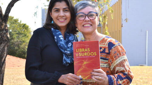 Duas mulheres sorridentes posam juntas ao ar livre segurando o livro "Libras e Surdos: Políticas, Linguagem e Inclusão". A mulher à esquerda veste blusa preta e cachecol azul com estampa floral; a da direita, de cabelos grisalhos curtos, usa óculos e uma blusa estampada colorida. Ambas olham para a câmera.