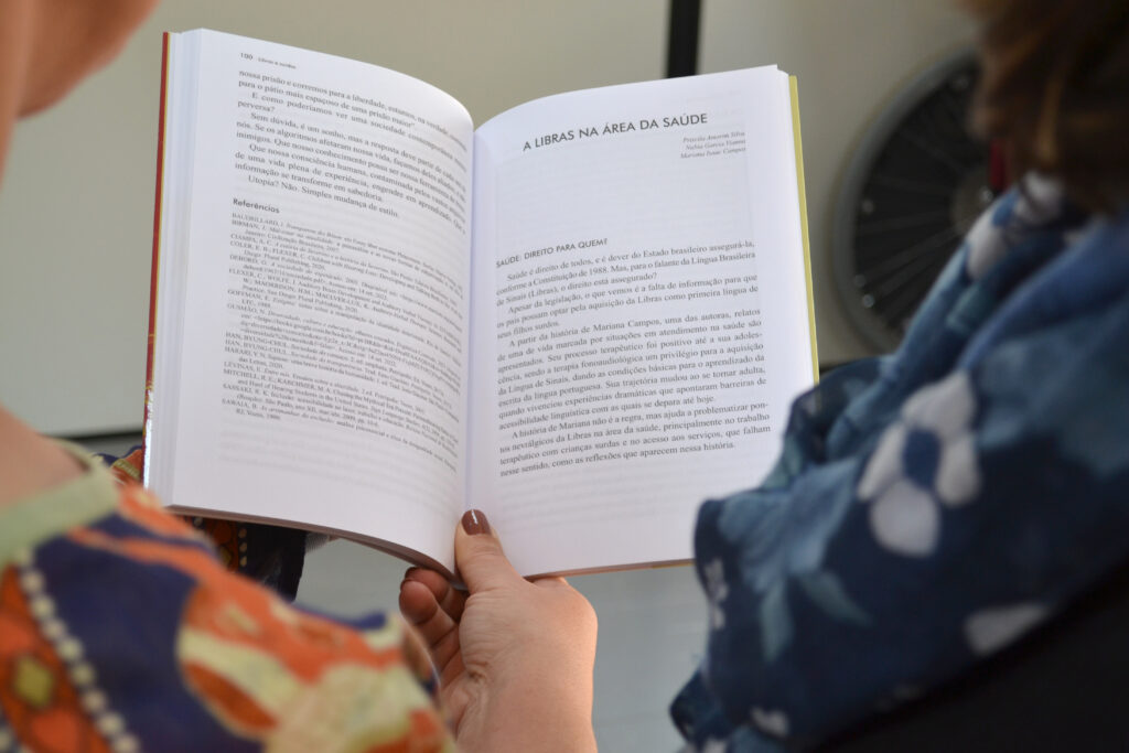 Duas mulheres leem juntas um exemplar aberto do livro "Libras e Surdos". A página exibida apresenta o capítulo intitulado “A Libras na área da saúde”. A imagem foca na leitura e nas mãos das leitoras, com o texto visível.