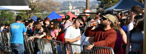 Fila extensa de pessoas jovens aguardando atrás de grades de contenção em área externa, sob sol forte. Muitas estão em pé, outras sentadas no chão. Um homem de boné bege e camiseta vermelha aparece em destaque à direita, bebendo água de uma garrafa. Ao fundo, tendas coloridas e placas de trânsito são visíveis. O clima é de calor e aglomeração.