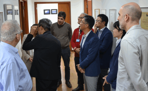 Grupo de oito pessoas em um corredor, observando uma parede com quadros e documentos emoldurados. Algumas pessoas estão olhando atentamente para um dos quadros, enquanto outras estão em diferentes posições, conversando ou observando os documentos. O ambiente é bem iluminado e os participantes estão vestidos de forma formal. O corredor tem portas de madeira e o piso é de madeira também. A cena parece ocorrer em um contexto de visita ou apresentação profissional.