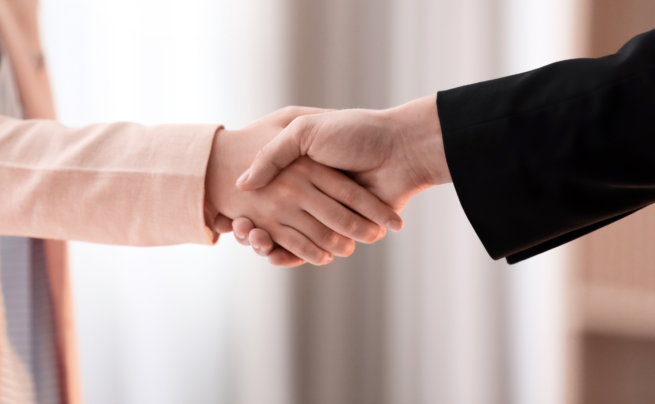 Texto alternativo para acessibilidade: Duas pessoas apertando as mãos, simbolizando um acordo ou cumprimento. Uma delas está vestindo um blazer rosa claro e a outra, um blazer preto. O fundo é desfocado, sugerindo um ambiente interno.