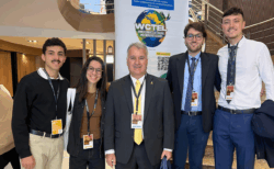 Cinco pessoas estão posando para uma foto em um evento. Ao centro, há um homem mais velho vestindo terno preto e gravata amarela. Ao seu redor, estão quatro jovens (dois de cada lado), todos com crachás de identificação pendurados no pescoço. À esquerda, um jovem de bigode usa suéter preto e calça clara, ao lado de uma jovem com cabelo solto, óculos e roupa casual. À direita, dois jovens de terno e gravata sorriem para a foto. Ao fundo, há uma escada e um banner com o logotipo do WCTEL (World Congress of Trauma and Emergency Leagues).