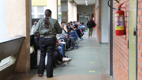 Pessoa de costas, usando jaqueta verde e calça preta, caminha por um corredor onde várias pessoas estão sentadas em bancos ao longo da parede, aparentando aguardar atendimento. O corredor tem piso verde, paredes de tijolos à direita, extintor de incêndio preso na parede e pilares revestidos de material rugoso à esquerda. Ao fundo, vê-se uma porta de vidro e mais pessoas sentadas.