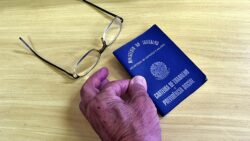 Pessoa idosa segura uma Carteira de Trabalho e Previdência Social azul, documento brasileiro utilizado para registro de vínculos empregatícios. Sobre a mesa de madeira clara, ao lado da carteira, está um par de óculos de grau com armação preta. Créditos da fotografia: Antoninho Perri/Unicamp
