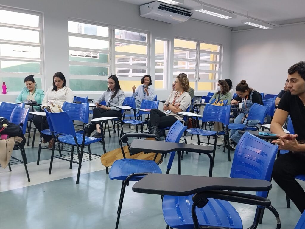 Participantes sentados em cadeiras azuis, a maioria voltada para frente. Uma mulher ao centro segura um microfone, enquanto outros escrevem ou observam. Ao fundo, grandes janelas deixam entrar luz natural.