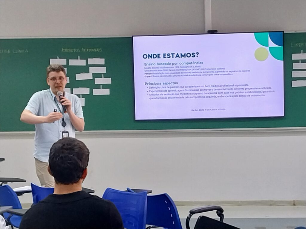 Homem com microfone em uma mão e apontador na outra apresenta conteúdo projetado na tela. O slide tem o título “ONDE ESTAMOS?” e trata de ensino baseado por competências, com tópicos e parágrafos em português. Algumas pessoas sentadas assistem.