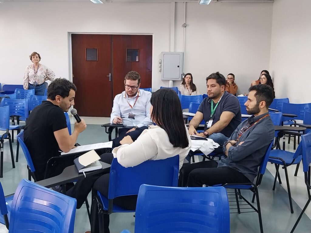 Grupo de seis pessoas reunidas em cadeiras azuis dispostas em círculo, debatendo. Um homem de camiseta preta fala ao microfone enquanto os demais escutam ou fazem anotações. Ao fundo, outros participantes e uma mulher em pé observando.