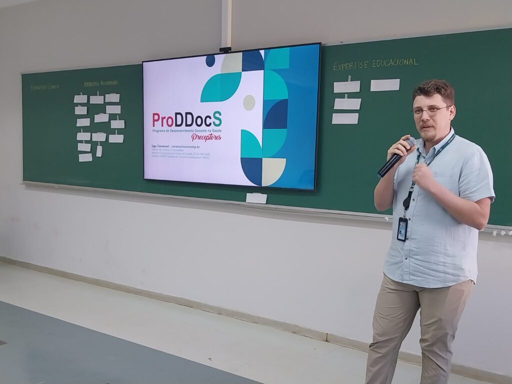 Homem de camisa clara apresenta em pé, segurando microfone. Ao lado dele, um telão exibe o título “ProDDocS Programa de Desenvolvimento Docente na Saúde – Preceptores”. Ao fundo, quadro verde com anotações e papéis colados.