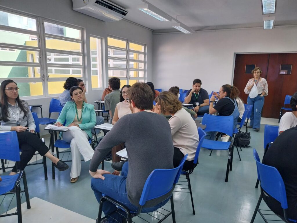 Sala de aula com grupos de pessoas conversando em pequenos círculos. Uma mulher em pé observa ao fundo. Algumas pessoas fazem anotações, outras discutem em duplas ou trios. Ao lado esquerdo, janelas grandes iluminam o ambiente.
