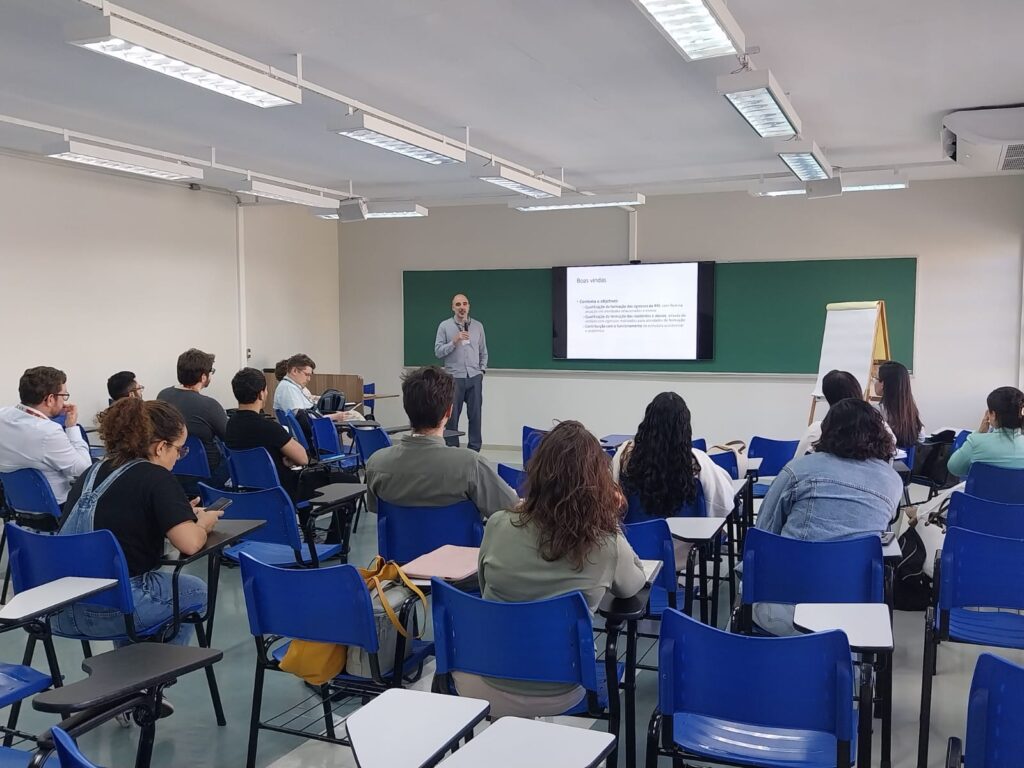 Homem em pé com microfone apresenta um slide projetado com o título “Boas vindas”. Participantes sentados observam a apresentação. Ao lado da tela, há um quadro verde e um flip chart branco.