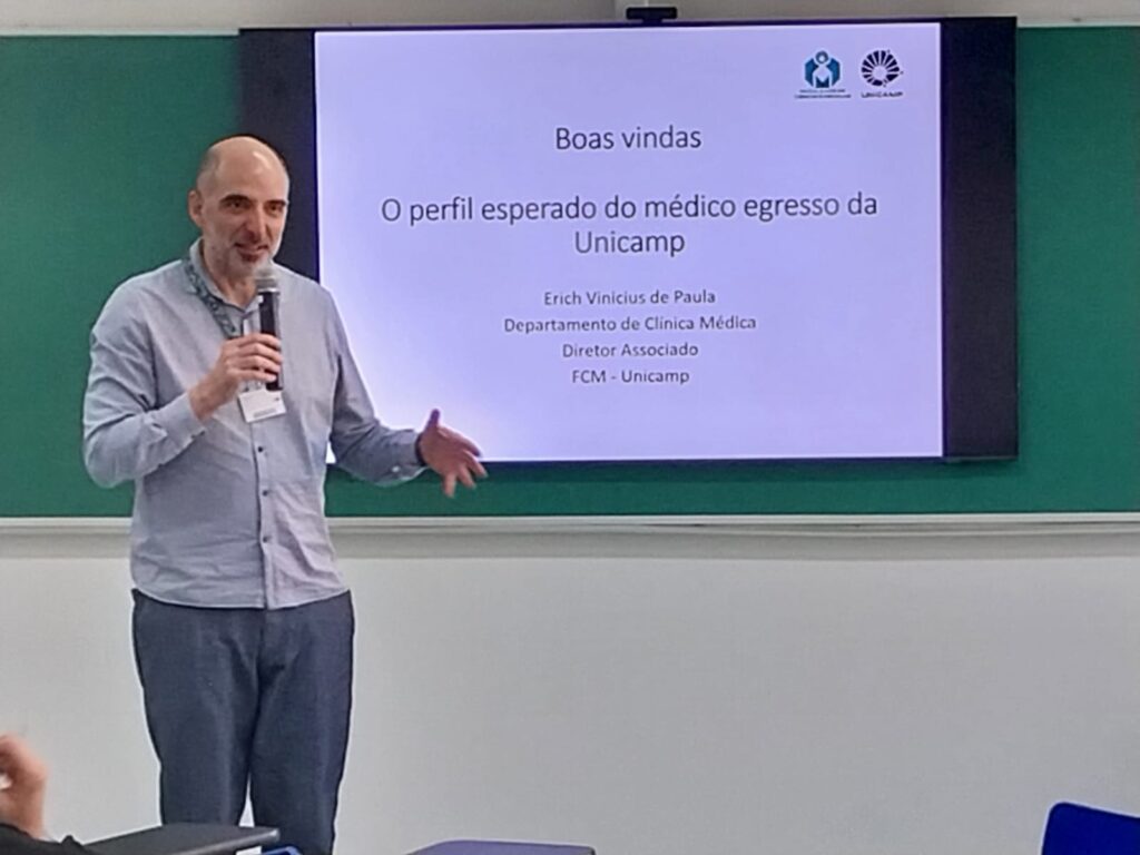 Um homem está de pé em uma sala de aula, segurando um microfone e falando ao público. Ele usa camisa azul clara e crachá no pescoço. Ao fundo, há uma tela com o slide projetado que diz: "Boas-vindas — O perfil esperado do médico egresso da Unicamp", com o nome Erich Vinicius de Paula, do Departamento de Clínica Médica, Diretor Associado da FCM - Unicamp. A tela está posicionada sobre um quadro verde.