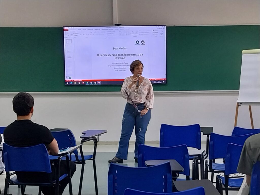 Uma mulher está de pé em uma sala de aula, segurando um microfone e falando ao público. Ela usa blusa clara estampada e calça jeans. Ao fundo, aparece a mesma apresentação projetada na tela com o título: "Boas-vindas — O perfil esperado do médico egresso da Unicamp", com o nome de Erich Vinicius de Paula. Há algumas pessoas sentadas em cadeiras azuis com mesas acopladas, e um quadro branco móvel está posicionado à direita da imagem.