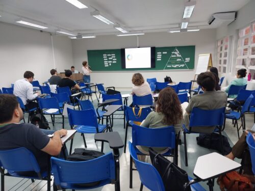 Sala de aula com cadeiras azuis parcialmente ocupadas por participantes atentos. Uma mulher em pé fala ao microfone perto de um telão que exibe um gráfico circular. Um homem está sentado ao fundo, próximo ao computador. Ao lado do telão, há um quadro verde com papéis colados em formato de pirâmide e outras anotações.