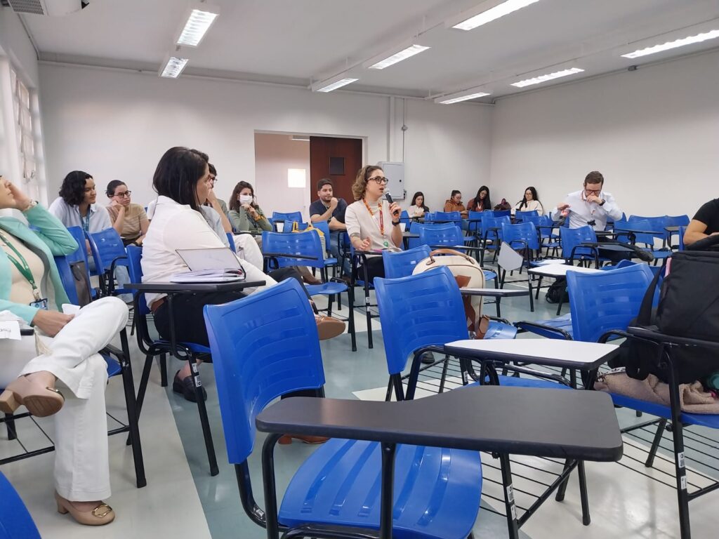 Vista lateral de uma sala com cadeiras azuis, onde participantes acompanham a apresentação. Uma mulher ao centro fala ao microfone, enquanto outros olham atentos ou fazem anotações. Algumas pessoas usam máscaras, sentadas nas últimas fileiras.