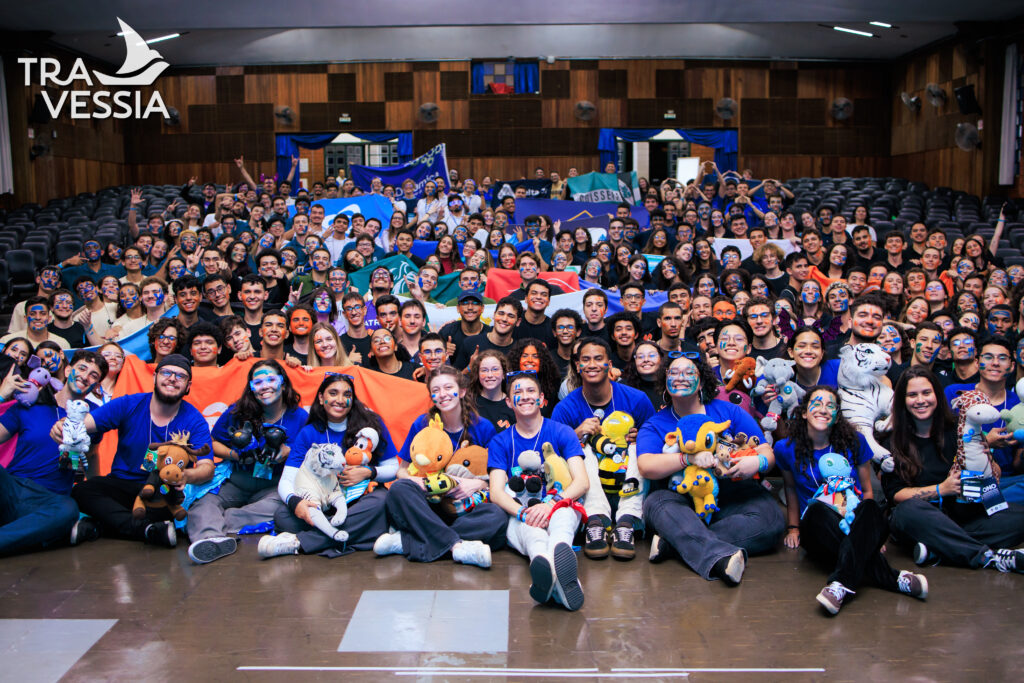 Grande grupo de jovens posa em auditório com bandeiras coloridas e rostos pintados, celebrando o evento Travessia. Todos sorriem e muitos seguram bichos de pelúcia.

