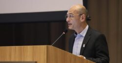 Homem calvo, de pele clara, usando óculos, camisa social azul-clara e paletó preto, discursa em um púlpito de madeira com microfone. No púlpito, há o logotipo e a inscrição “Organização Pan-Americana da Saúde”. Ao fundo, cortina escura e parte de uma tela de projeção.