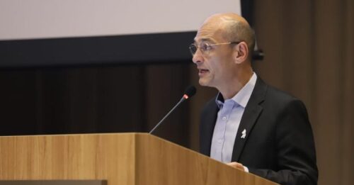 Homem calvo, de pele clara, usando óculos, camisa social azul-clara e paletó preto, discursa em um púlpito de madeira com microfone. No púlpito, há o logotipo e a inscrição “Organização Pan-Americana da Saúde”. Ao fundo, cortina escura e parte de uma tela de projeção.