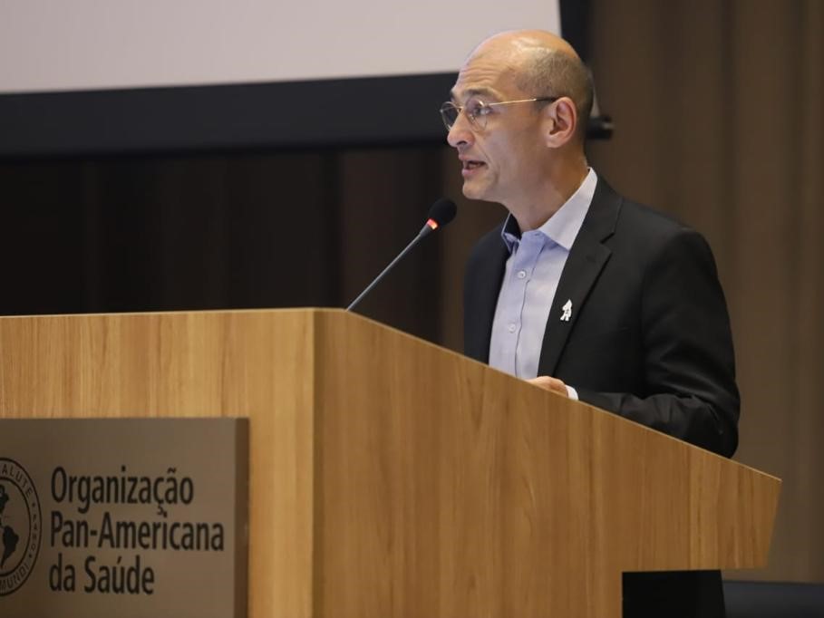 Homem calvo, de pele clara, usando óculos, camisa social azul-clara e paletó preto, discursa em um púlpito de madeira com microfone. No púlpito, há o logotipo e a inscrição “Organização Pan-Americana da Saúde”. Ao fundo, cortina escura e parte de uma tela de projeção.