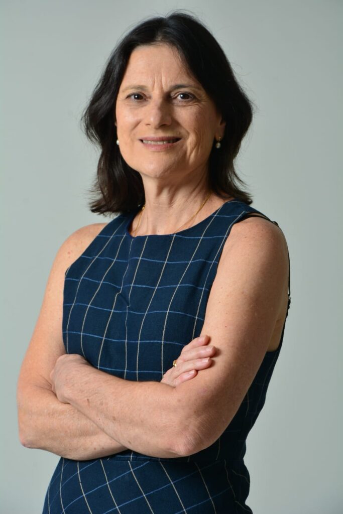 A professora Maria Francisca Colella dos Santos, coordenadora do curso de graduação em Fonoaudiologia da Faculdade de Ciências Médicas da Unicamp, está de pé, com os braços cruzados e sorrindo levemente. Ela veste um vestido azul-marinho com estampa de listras formando quadros. Ao fundo, há uma parede cinza-clara. Foto: Mário Moreira.