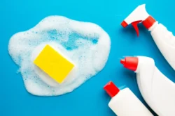Esponja amarela sobre espuma branca de sabão em um fundo azul, ao lado de três frascos brancos de produtos de limpeza com tampas vermelhas, incluindo um borrifador.