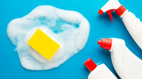 Esponja amarela sobre espuma branca de sabão em um fundo azul, ao lado de três frascos brancos de produtos de limpeza com tampas vermelhas, incluindo um borrifador.