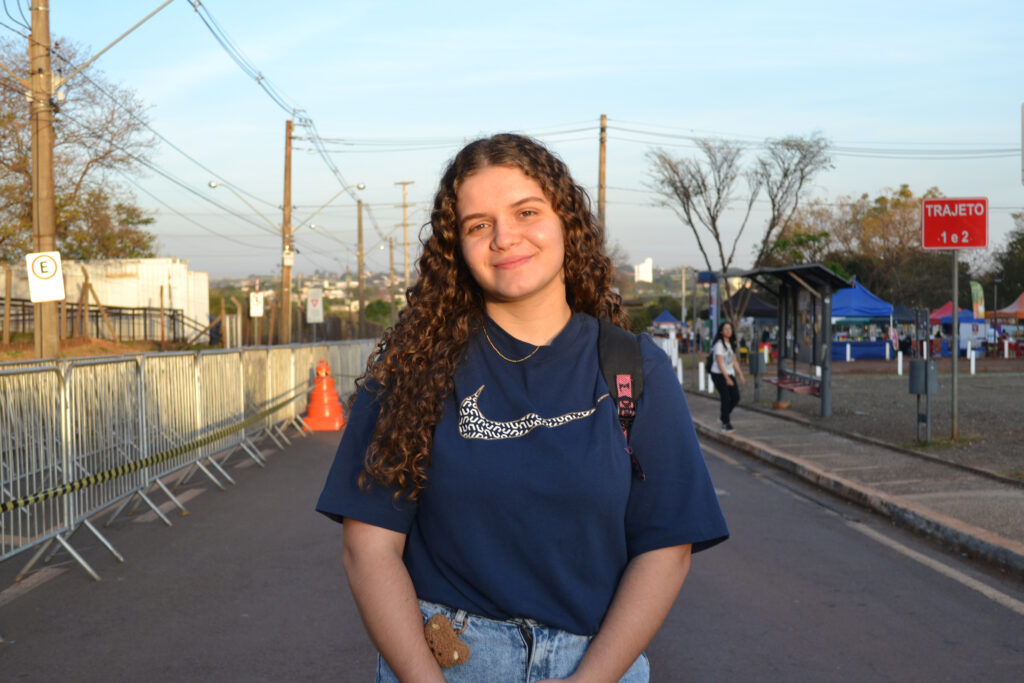 Uma jovem de cabelos cacheados castanhos, usando camiseta azul marinho com estampa clara e mochila preta, sorri para a câmera em uma rua com gradis metálicos e barracas coloridas ao fundo.