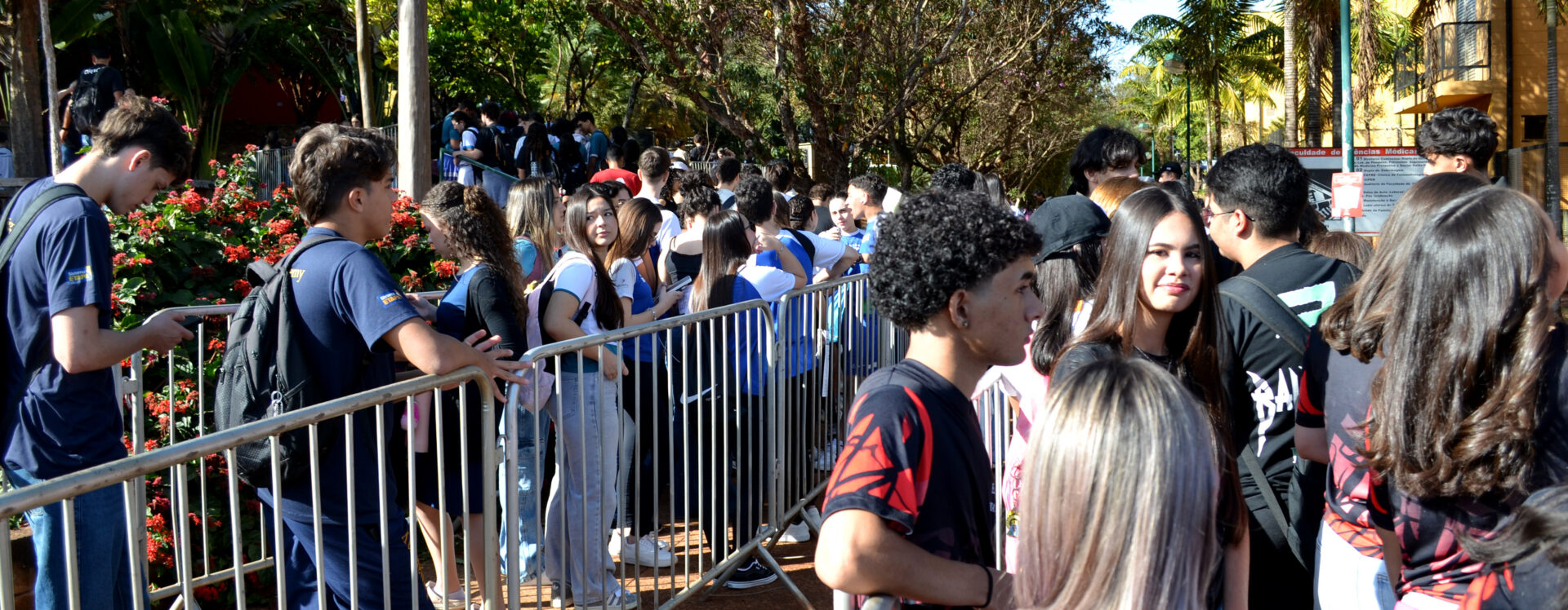 Estudantes aguardam em fila organizada por grades metálicas em área externa arborizada, durante evento acadêmico.