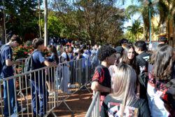 Estudantes aguardam em fila organizada por grades metálicas em área externa arborizada, durante evento acadêmico.