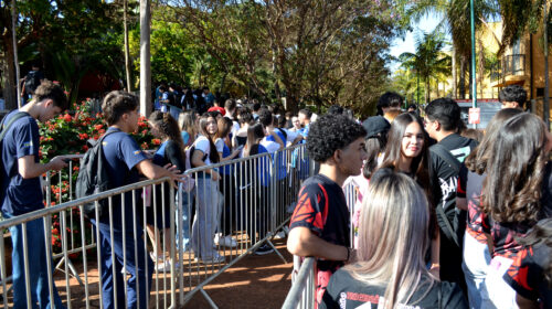 Estudantes aguardam em fila organizada por grades metálicas em área externa arborizada, durante evento acadêmico.