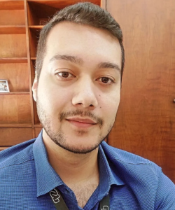 Homem de barba rala e cabelo curto escuro, vestindo uma camisa azul e crachá preto, tira uma selfie em um ambiente de escritório com estante de madeira ao fundo.