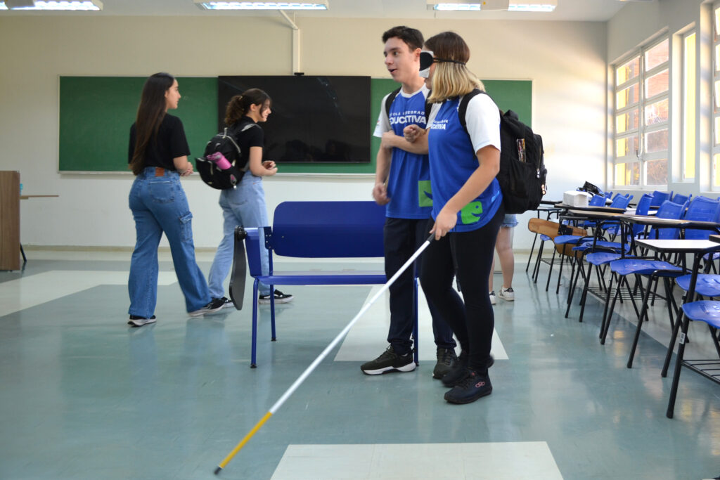 Estudantes em sala de aula participam de dinâmica sobre acessibilidade; uma jovem com venda nos olhos e bengala é guiada por um colega, enquanto outros observam.