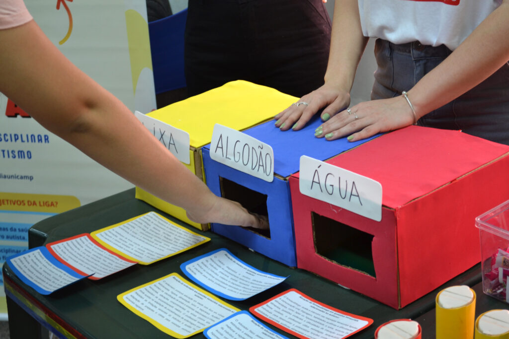 Participantes interagem com caixas coloridas (amarela, azul e vermelha), identificadas com as palavras “Lixa”, “Algodão” e “Água”, explorando texturas em uma atividade sensorial.