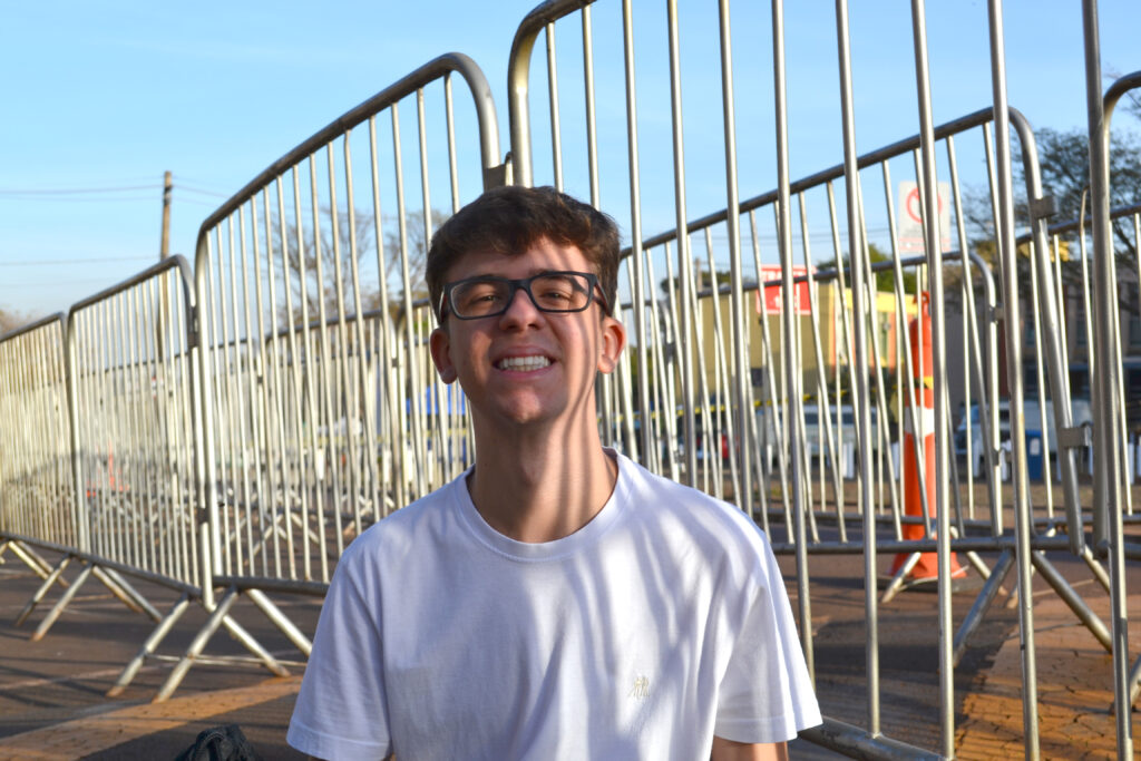 Um jovem de óculos e camiseta branca sorri amplamente para a câmera, sentado próximo a gradis metálicos em uma área externa ensolarada.