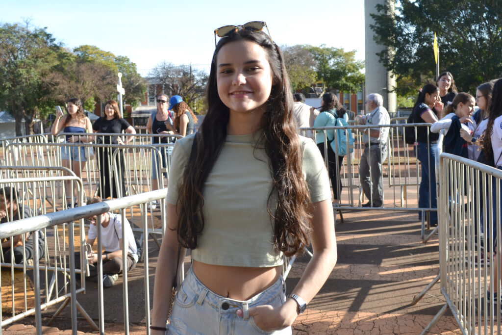 Uma jovem de cabelos longos e soltos, usando camiseta verde clara curta e shorts jeans, sorri para a câmera em meio a gradis, com várias pessoas ao fundo em um ambiente externo ensolarado.
