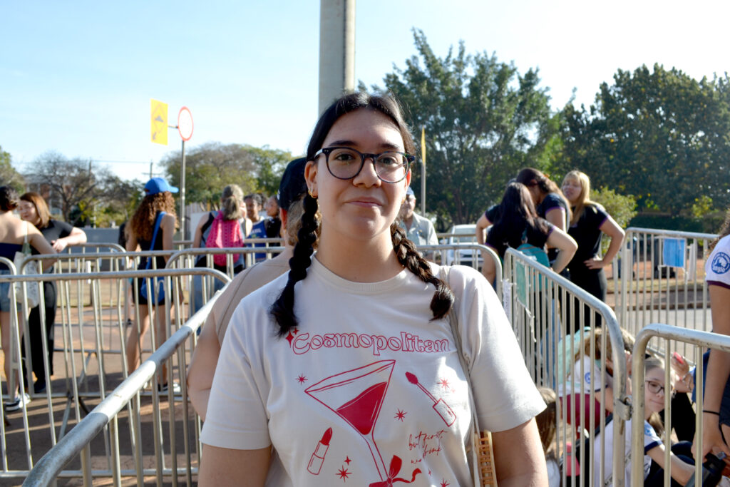 Uma jovem de óculos e tranças, vestindo camiseta clara com estampa vermelha da palavra “Cosmopolitan” e desenhos de batom e taça, olha para a câmera em meio a gradis, com outras pessoas ao fundo.