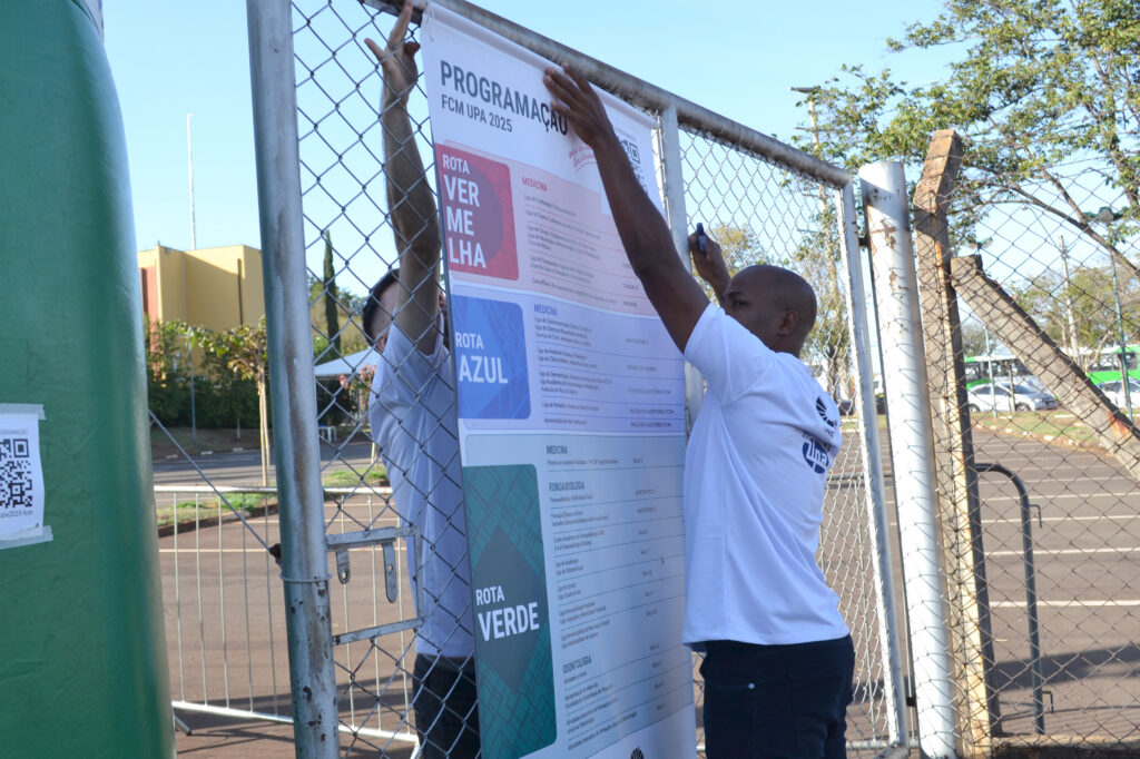 Duas pessoas fixam em uma grade metálica um grande banner com a programação da FCM UPA 2025. O banner exibe rotas identificadas por cores: vermelha, azul e verde, relacionadas às atividades dos cursos da área da saúde.