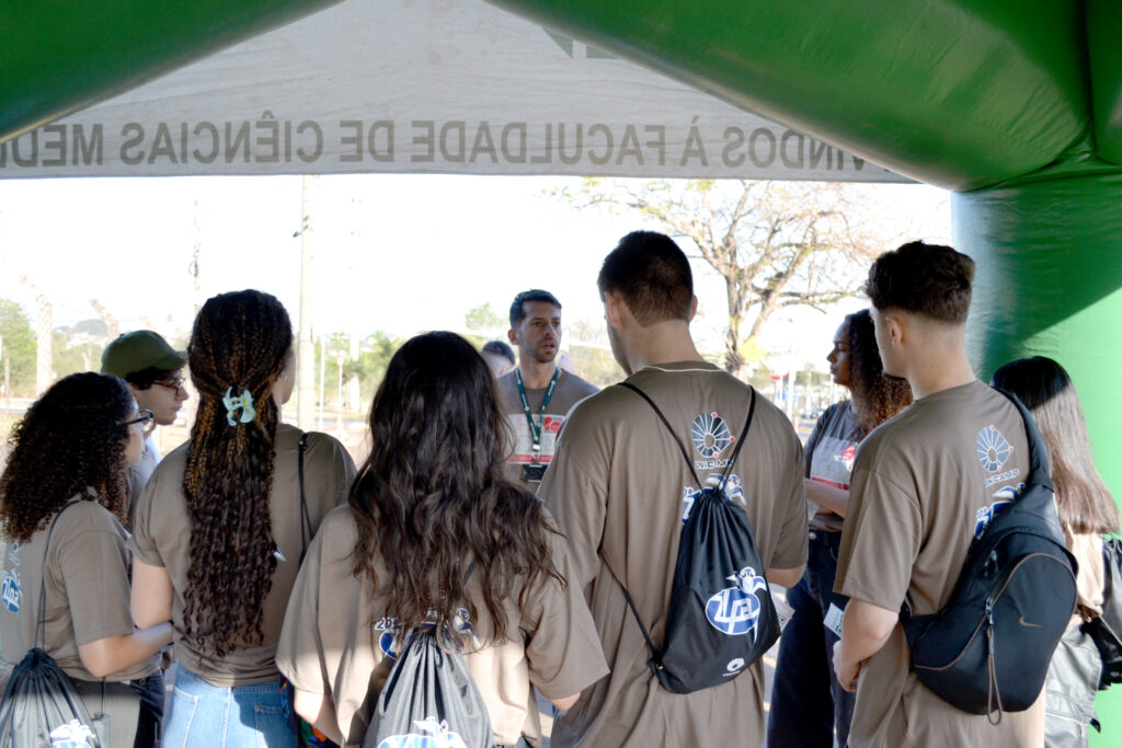 Grupo de jovens veste camisetas marrons com logotipo da Unicamp nas costas e ouve atentamente a explicação de um orientador, embaixo de uma tenda verde com a inscrição “Faculdade de Ciências Médicas”.