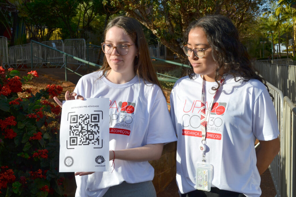 Duas jovens, usando camisetas brancas do evento UPA 2025, seguram um cartaz com QR Code impresso, que contém instruções pré-atividade. Ao fundo, há flores vermelhas e uma área com grades de contenção.