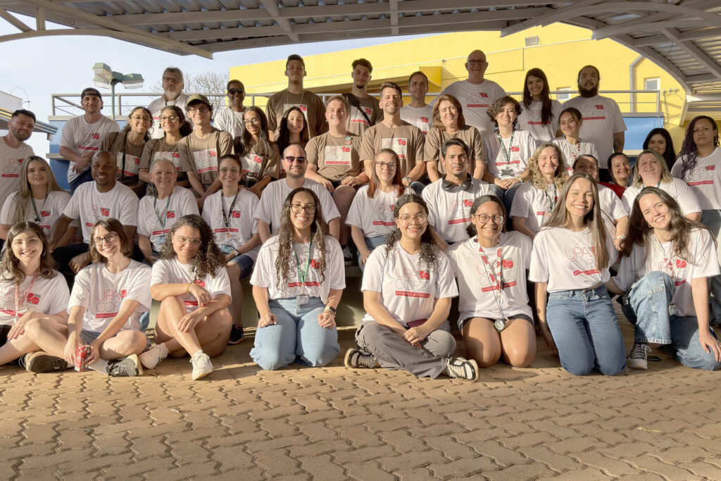 Foto em grupo de participantes e organizadores da FCM UPA 2025, reunidos em arquibancada ao ar livre. Pessoas usam camisetas brancas e marrons personalizadas do evento, posando para a foto coletiva e sorrindo.
