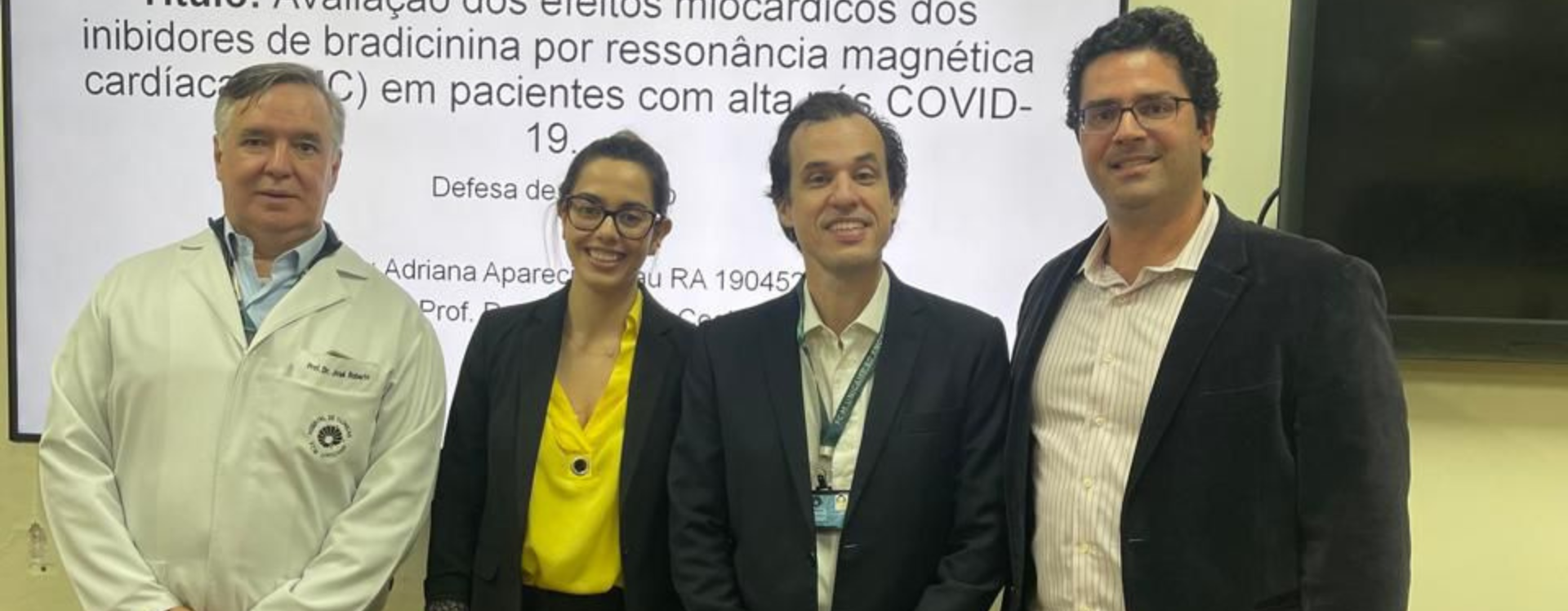 Quatro pessoas posam sorridentes em frente a uma tela de apresentação na Unicamp. A tela mostra o título de um trabalho acadêmico: “Avaliação dos efeitos miocárdicos dos inibidores de bradicinina por ressonância magnética cardíaca (RMC) em pacientes com alta de COVID-19”. À esquerda, um homem usa jaleco branco. Ao lado, uma mulher veste blazer preto e blusa amarela. À direita dela, um homem de terno escuro e crachá, e outro homem com blazer preto, camisa listrada e jeans.