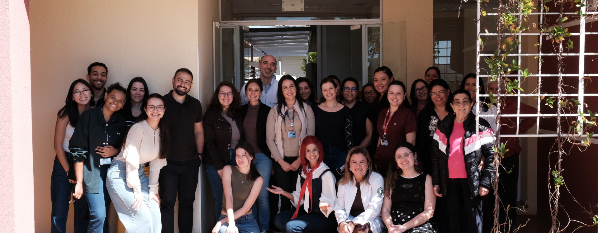 Grupo de cerca de 25 pessoas, em sua maioria mulheres, posando sorridentes para foto em frente a um prédio com porta de vidro. Algumas estão agachadas na frente e outras em pé atrás, em clima descontraído e de confraternização