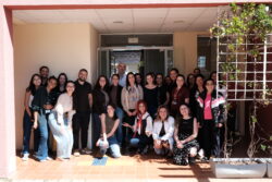 Grupo de cerca de 25 pessoas, em sua maioria mulheres, posando sorridentes para foto em frente a um prédio com porta de vidro. Algumas estão agachadas na frente e outras em pé atrás, em clima descontraído e de confraternização
