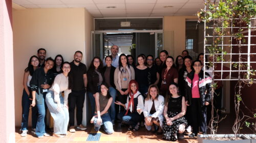 Grupo de cerca de 25 pessoas, em sua maioria mulheres, posando sorridentes para foto em frente a um prédio com porta de vidro. Algumas estão agachadas na frente e outras em pé atrás, em clima descontraído e de confraternização