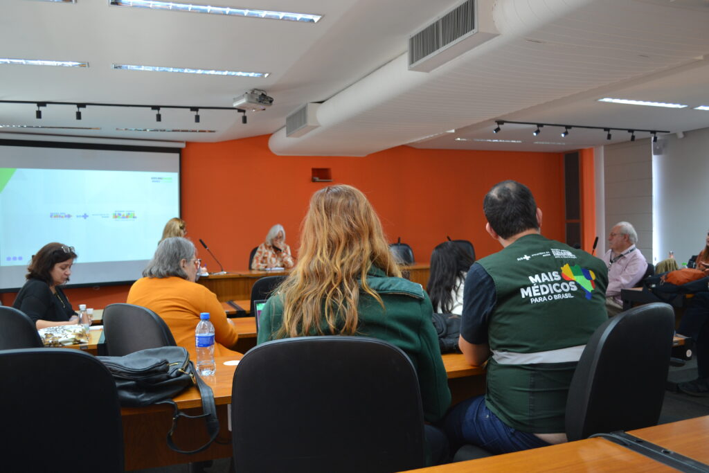 Reunião do Programa Mais Médicos realizada na FCM, com foco na ampliação e qualificação da Atenção Básica. Participantes estão sentados em volta de uma mesa em sala de conferência com parede laranja ao fundo. Duas pessoas vestem coletes verdes com o logotipo do programa.